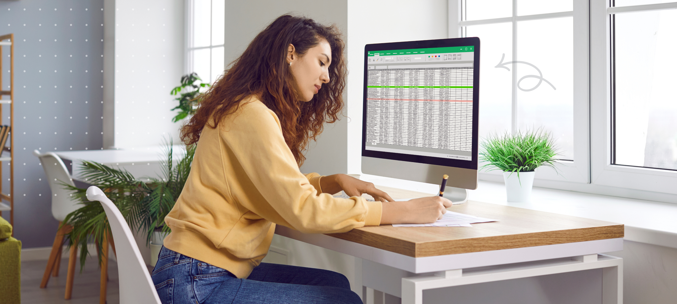 Image of woman sitting at desk, writing on paper in front of a monitor with a speadsheet on the screen